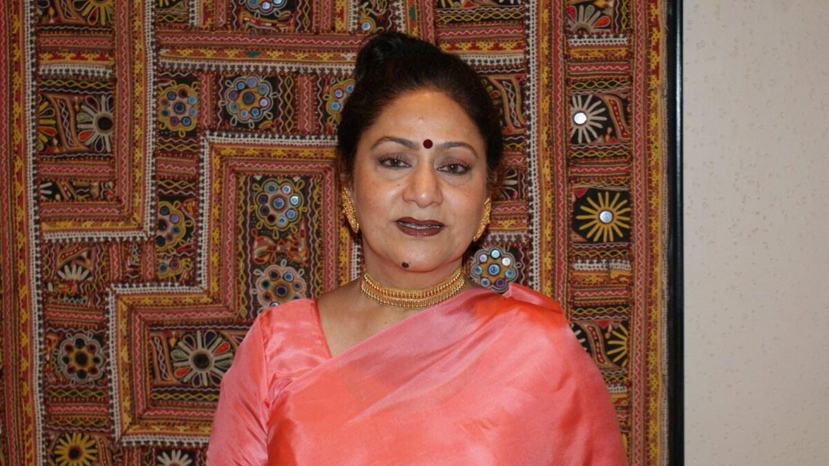 Aruna Irani in a pink saree standing in front of an embroidered wall hanging