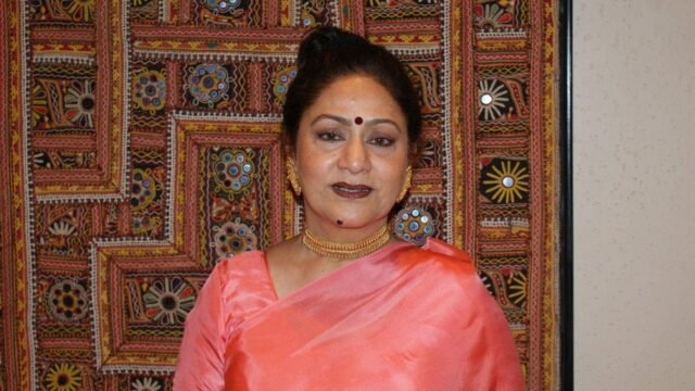 Aruna Irani in a pink saree standing in front of an embroidered wall hanging