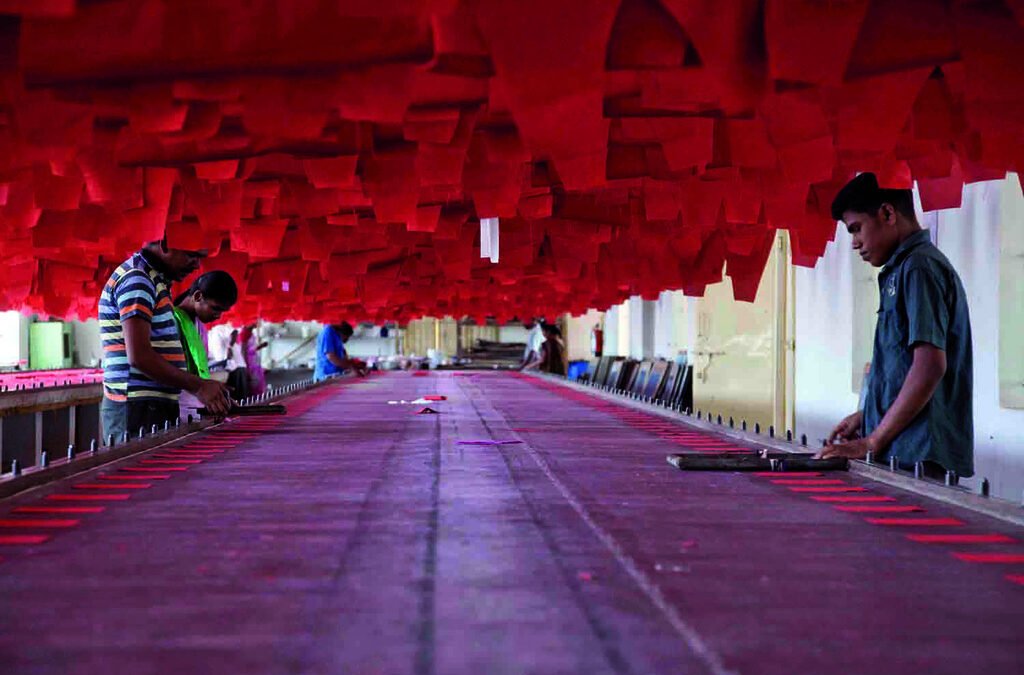 Workers at tables under rows of red fabric in an Indian garment factory