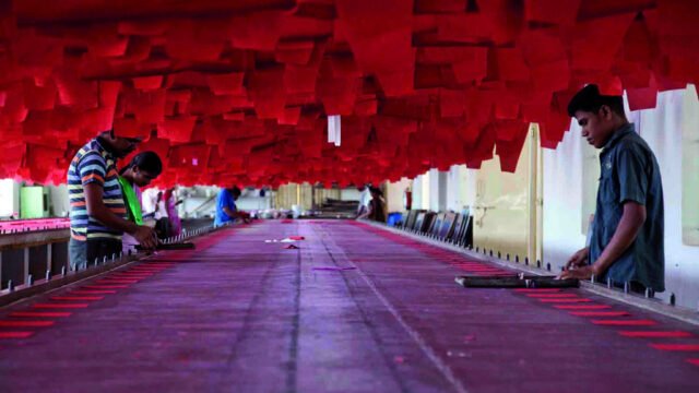Workers at tables under rows of red fabric in an Indian garment factory